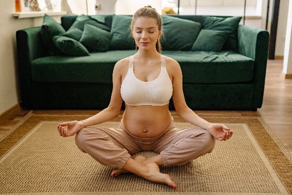 Une femme enceinte faisant du yoga, assise en tailleur, les yeux fermés, en brassière de sport et pantalon jogging, en pleine préparation à la naissance.