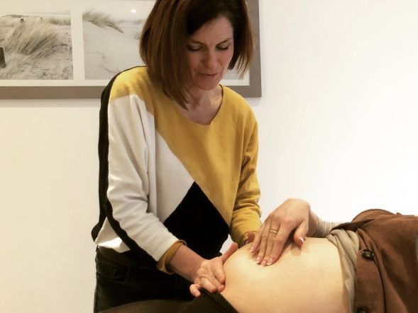 Sage-femme faisant un suivi de grossese à Bruxelles, en train d'ausculter le ventre d'une femme enceinte allongée.